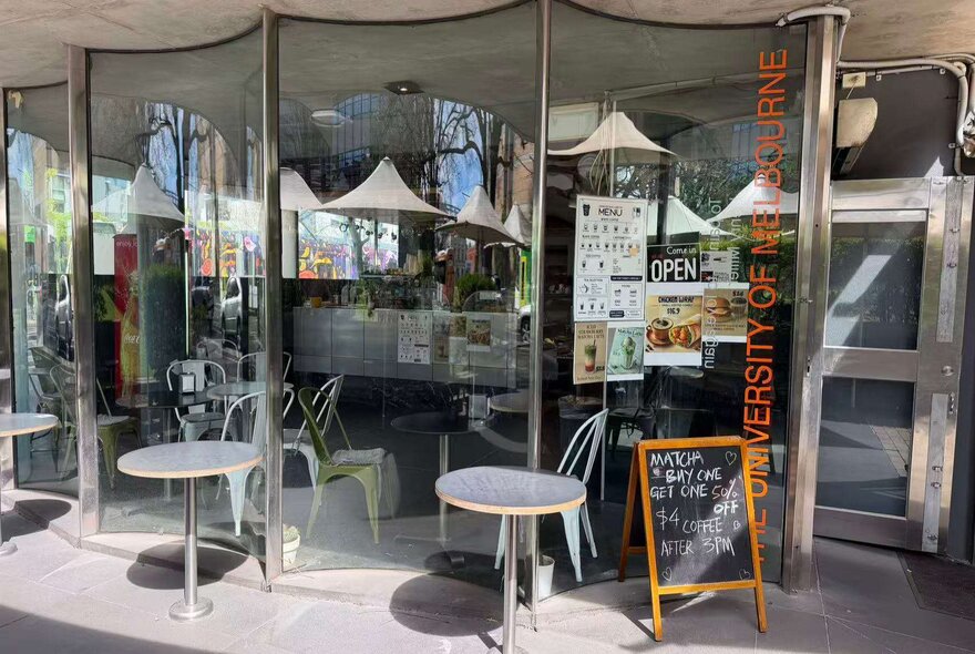 A university cafe with tables outside the curved glass windows with a menu board at the entrance.