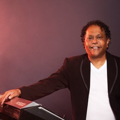 Soul musician Dinash Daniel resting his hand on a piano keyboard as he poses for a portrait photo in a studio setting.