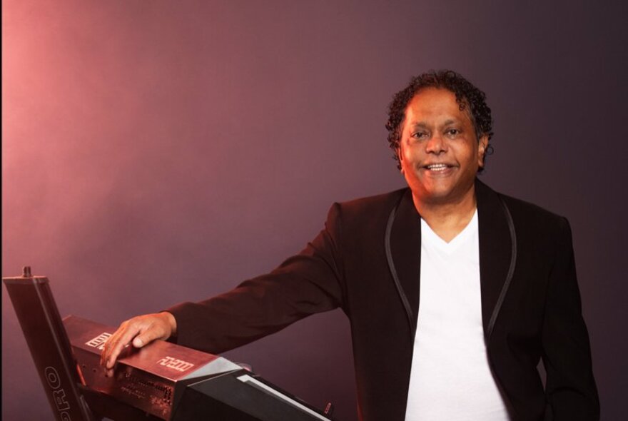 Soul musician Dinash Daniel resting his hand on a piano keyboard as he poses for a portrait photo in a studio setting.