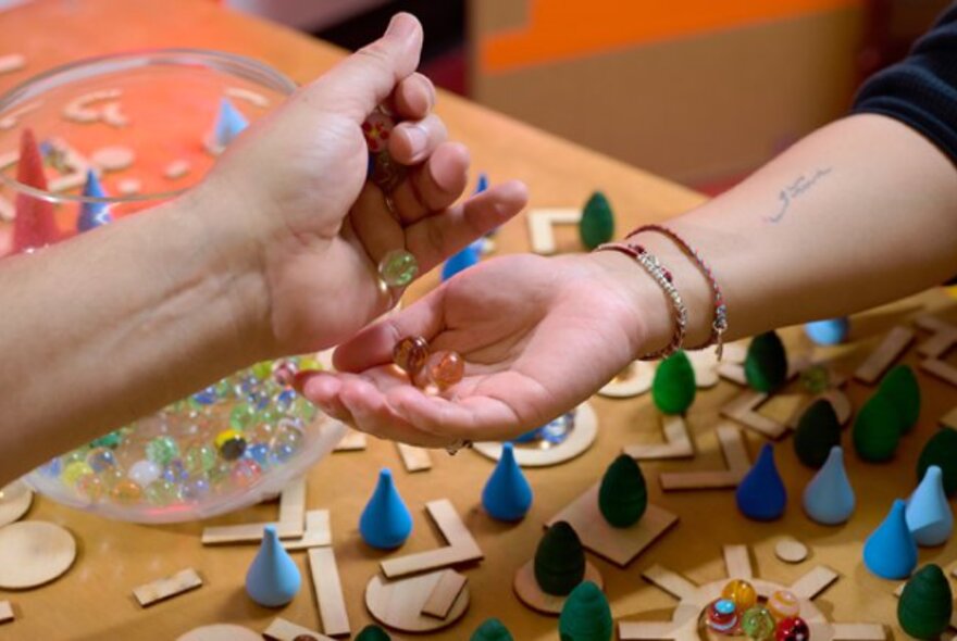 Two people's forearms leaning over opposite sides of a table, one hand pouring marbles into the other person's hand, with small puzzle and game pieces on the table below.