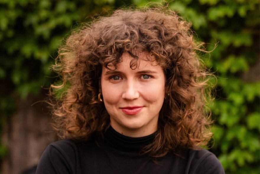 A woman with voluminous, curly brown hair, a slight smile, wearing a black top, standing outdoors in front of greenery.
