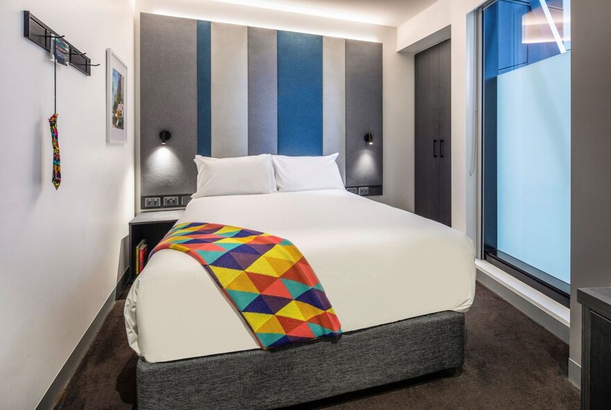 A contemporary hotel room at the ibis Styles Melbourne Southbank featuring a queen-sized bed with a white duvet and a colourful, patterned throw blanket.
