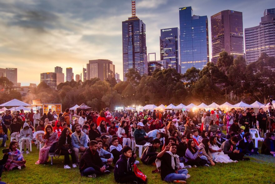 Crowds at Birrarung Marr at sunset with city buildings in the background and stalls on the grass.