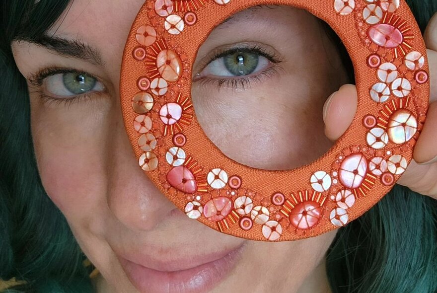 A smiling woman holding a hand-made circular artistic creation to her face and looking through it.