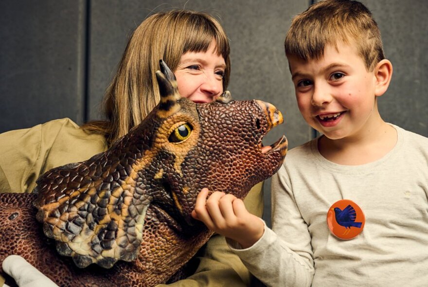 A child smiling at the camera and touching the cheek of  a dinosaur puppet being controlled by a woman who holds the puppet in her arms.