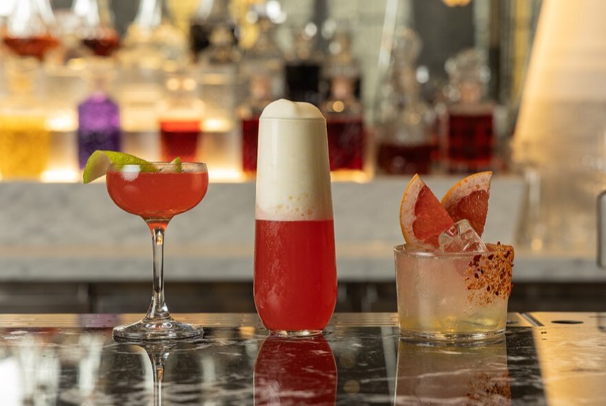 Three pink-toned cocktails lined up on a bar, each in a different shaped glass.
