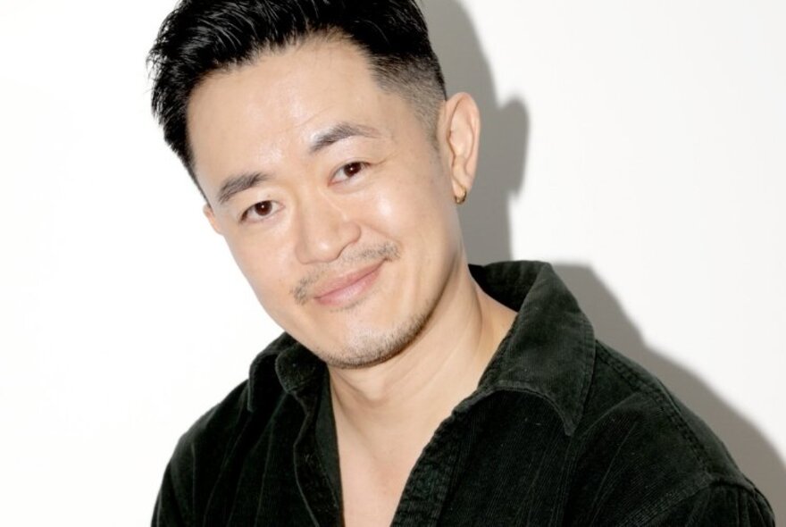 Playwright and author Benjamin Law looking directly at the viewer with a slight smile on his face, wearing a black shirt and standing in front of a white backdrop.