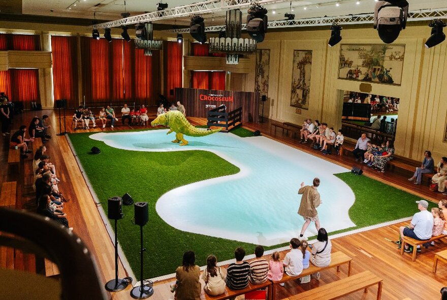 A crocodile character in a theatrical pool and grass area, children seated on benches looking on.