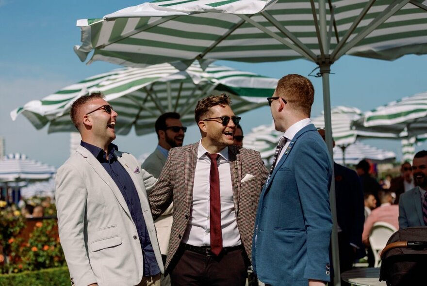 Three men in jackets and ties laughing under a stripy umbrella