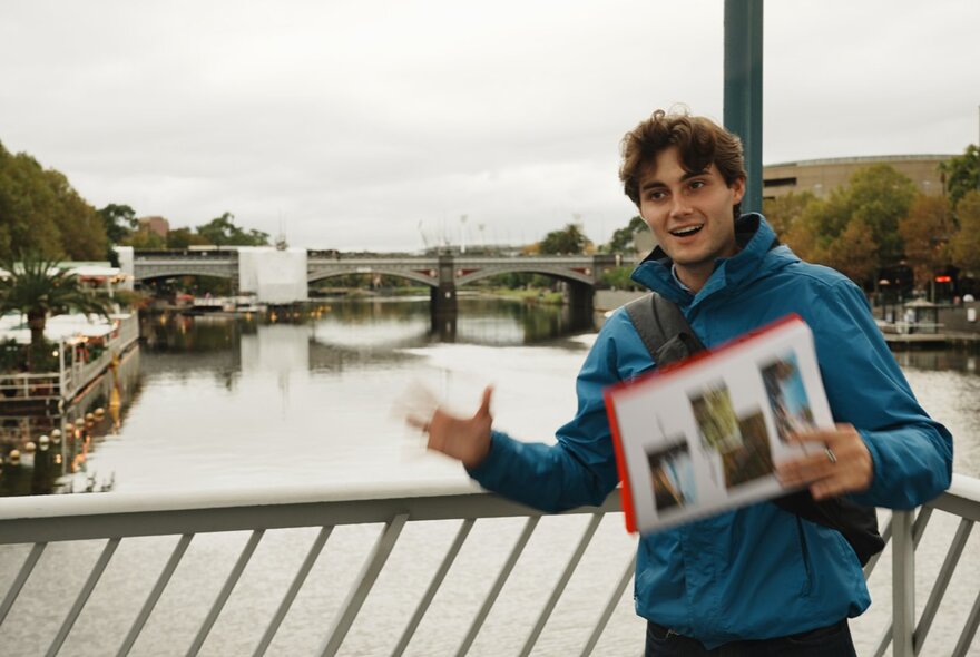 Young man holding an illustrated flyer, standing on a bridge over the Yarra.