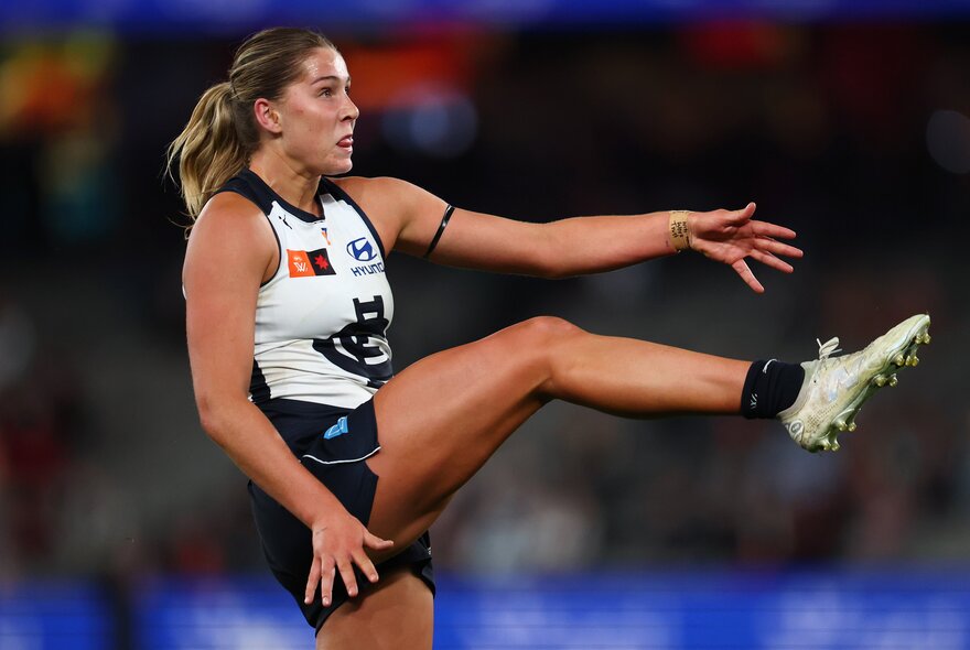 A Carlton AFLW player with her leg raised high in mid-air, having just kicked the football. 