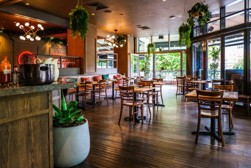 Restaurant interior with wooden floors, tables and chairs, windows looking onto greenery and moody lighting.