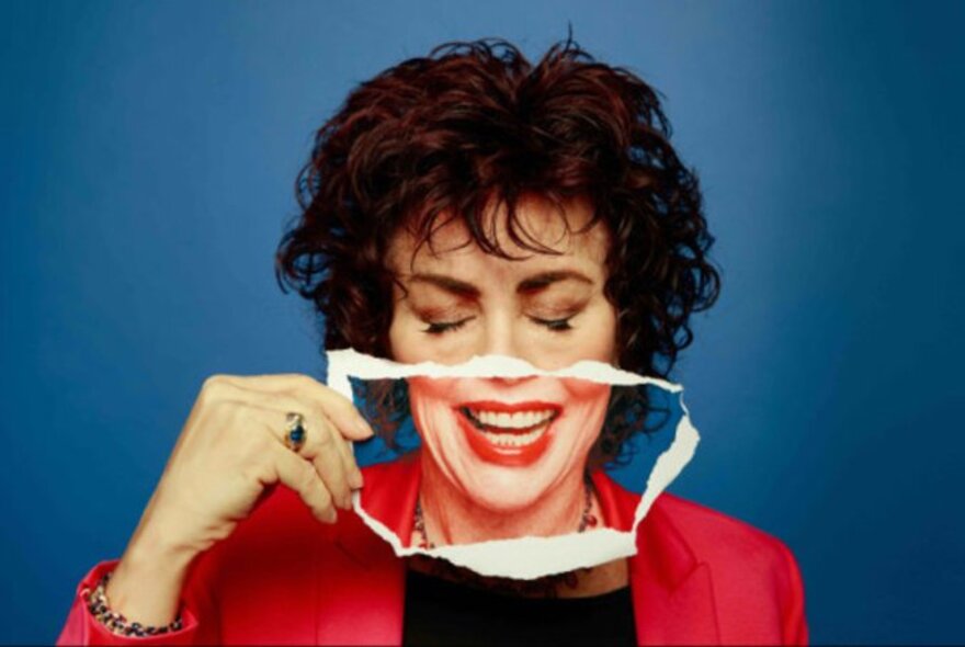 Comedian Ruby Wax holding up at cut out of her mouth laughing over her mouth. 