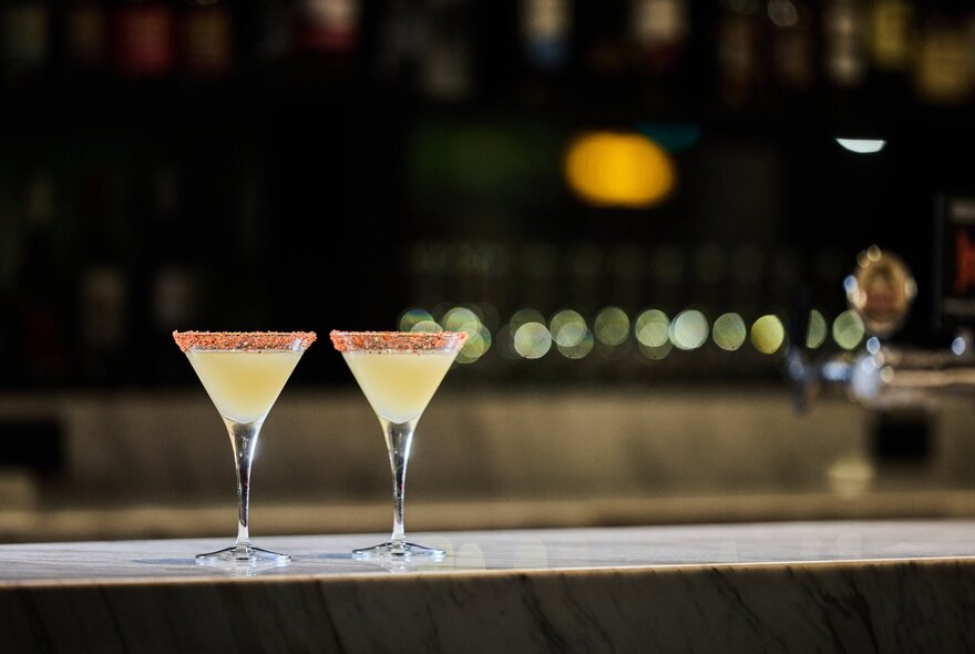 Two martinis on a bar counter with twinkling lights behind.