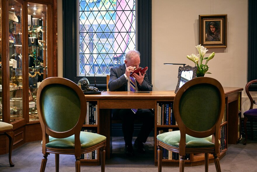 A formal room with a man sitting at a desk, two empty velvet chairs in front of him, while he peers through an eyeglass at a small object in his hand. 