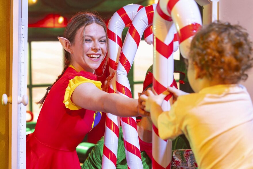 An actor dressed as a Christmas elf with pointed ears smiles while handing a large, inflatable candy cane to a young toddler.