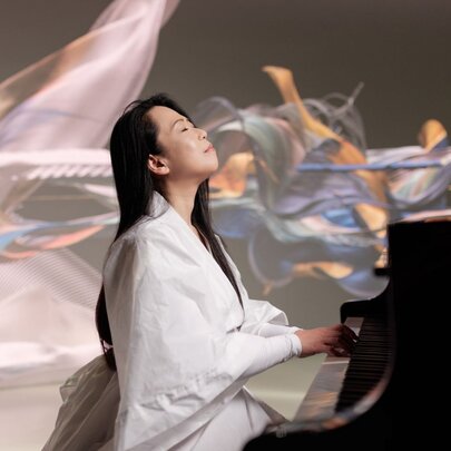 A pianist in a white road plays the piano while looking upwards, holographic shapes flowing in the background. 