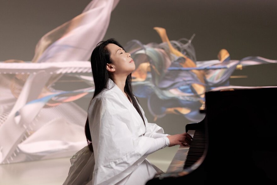 A pianist in a white road plays the piano while looking upwards, holographic shapes flowing in the background. 