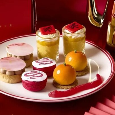 A selection of Lunar New Year themed high tea desserts, presented on a white, gold-rimmed plate with a red background.