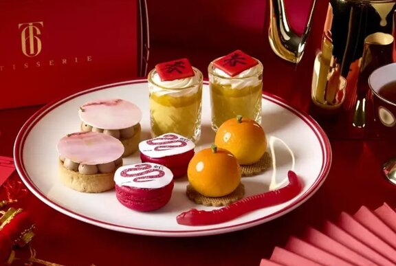 A selection of Lunar New Year themed high tea desserts, presented on a white, gold-rimmed plate with a red background.