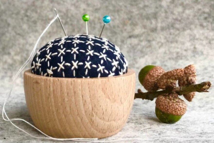 A pin cushion with a wooden base and pins sticking out the top and a gumnut. 