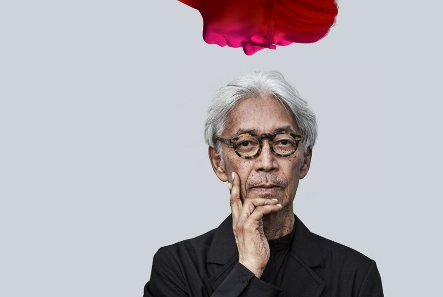 The late Sakamoto with his hand to his chin, with grey hair and tortoiseshell spectacles, a red face looking down on him from above.