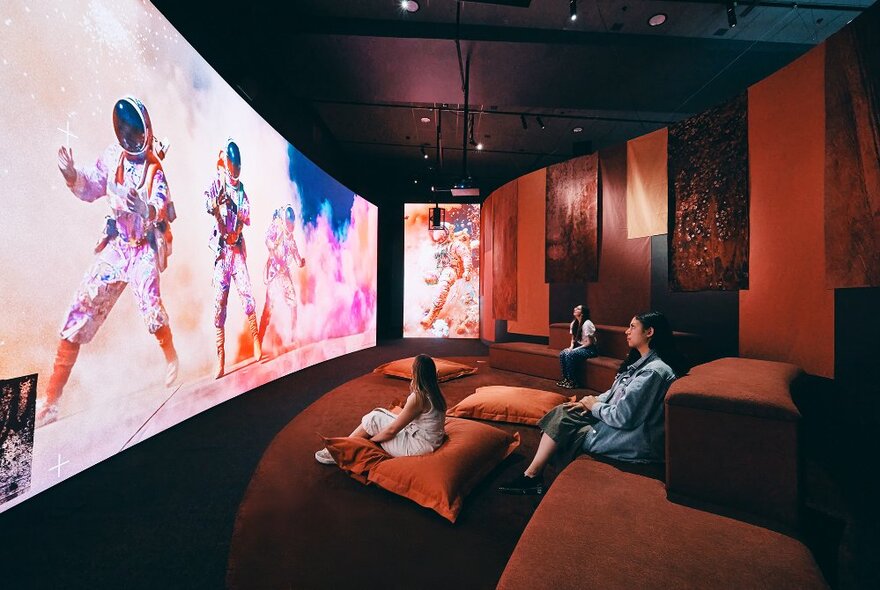 People seated in a relaxed viewing space with cushions, watching astronauts on a huge screen.