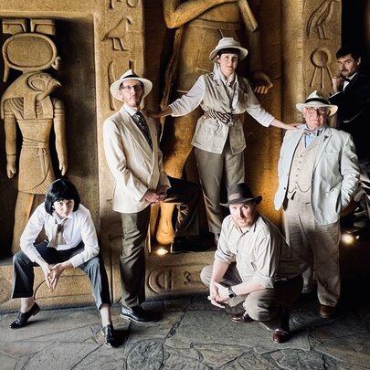 Five people dressed in 1920s explorer costumes standing in front of a wall of Egyptian hieryoglyphics.