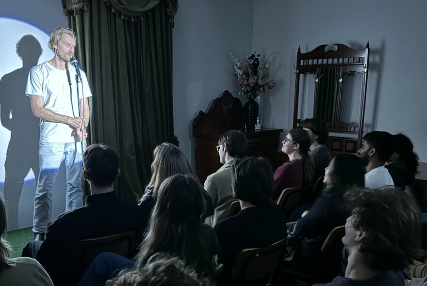 A stand up comedian on a small stage performing to a seated audience.