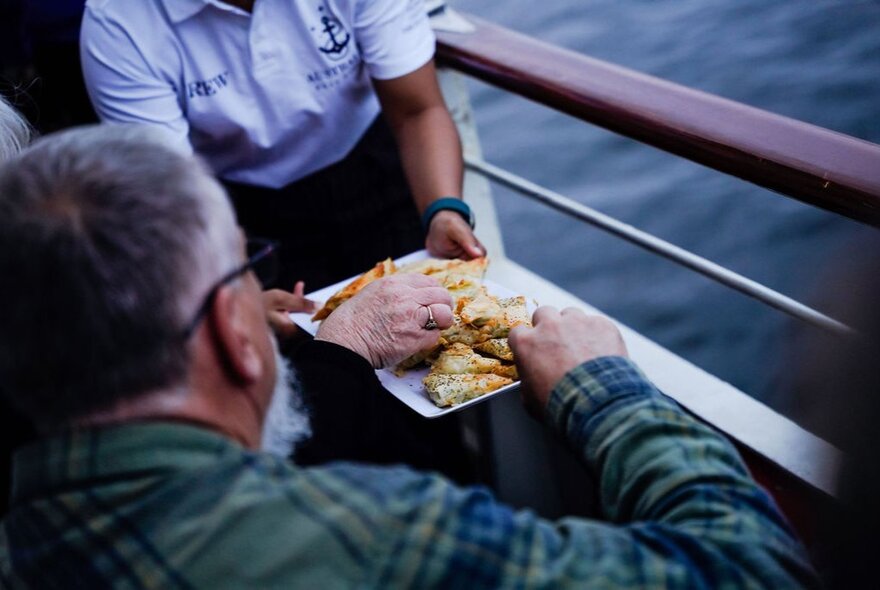 A man on a boat eating food from a plate being held by a crew member. 