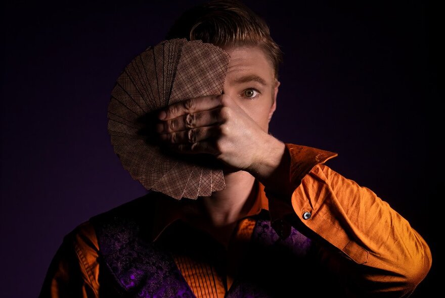 A performer in an orange shirt and purple vest, holding a fanned-out deck of cards over half his face.