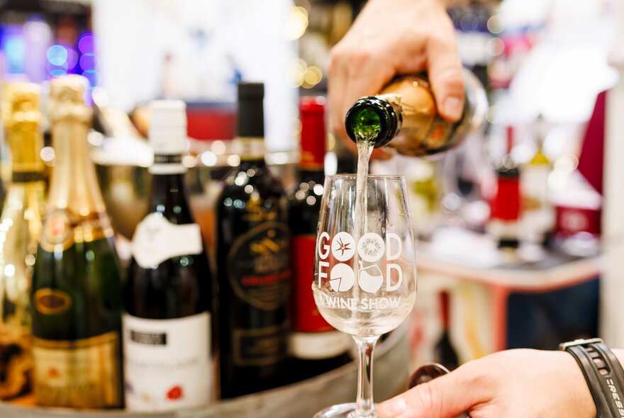 Hand pouring wine into a branded glass with bottles in the background.