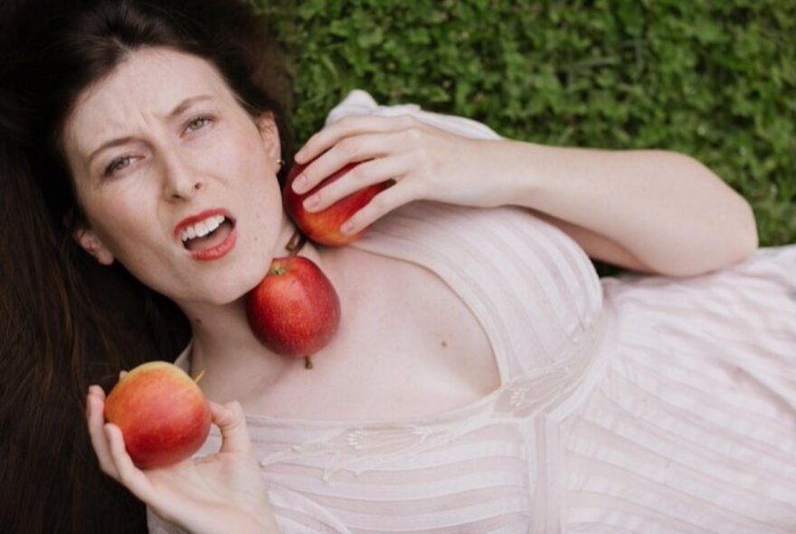 Woman lying on her back on the ground holding three apples.