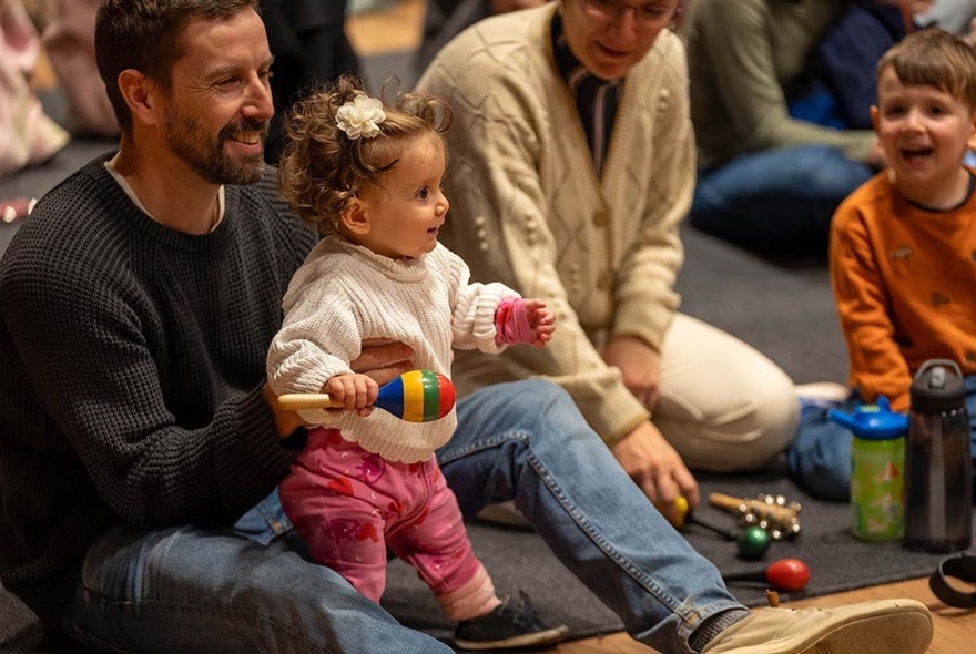 A toddler sitting on a man's knees seated on the floor with people looking on.