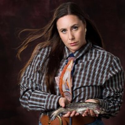 Comedian Kate Dolan, a woman with long brown hair, wearing a plaid shirt, an orange patterned tie, holding a small trout in her hands against a dark brown background.