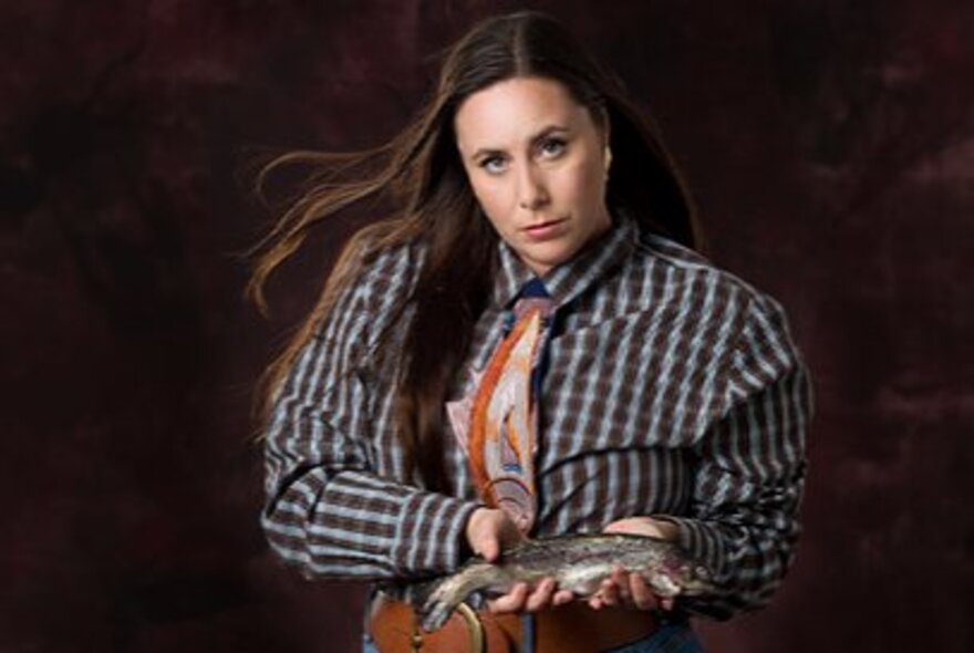 Comedian Kate Dolan, a woman with long brown hair, wearing a plaid shirt, an orange patterned tie, holding a small trout in her hands against a dark brown background.