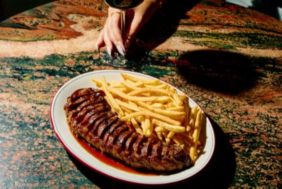 A Wagyu rump steak and chips on a plate sliced with a hand holding a glass of wine.