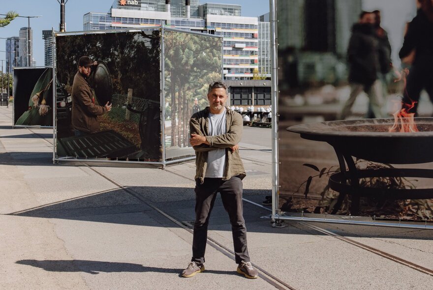 A man standingn in front of large scale artworks depicting smoking ceremonies, outdoors at Melbourne's Docklands. 