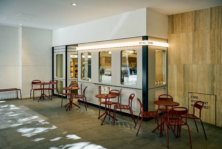 Exterior of cafe tucked into the corner of a wall, with small red tables and chairs out the front.