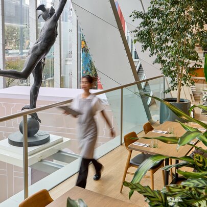 Blurred motion image of a waiter in an apron walking through a glass atrium area with a large statue, empty tables and greenery around. 