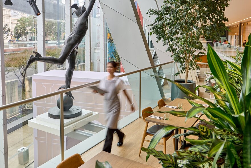 Blurred motion image of a waiter in an apron walking through a glass atrium area with a large statue, empty tables and greenery around. 