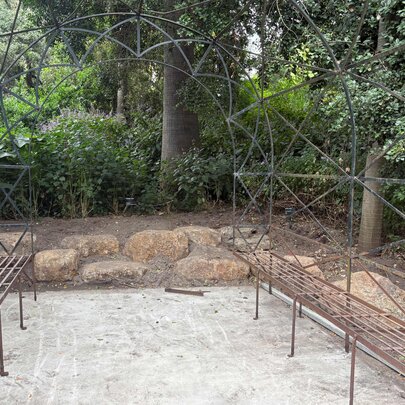A metal garden arch or arbour with a weathered rusted finish, surrounded by plantings and trees in the background.