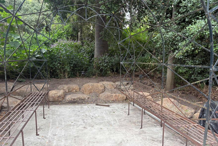 A metal garden arch or arbour with a weathered rusted finish, surrounded by plantings and trees in the background.