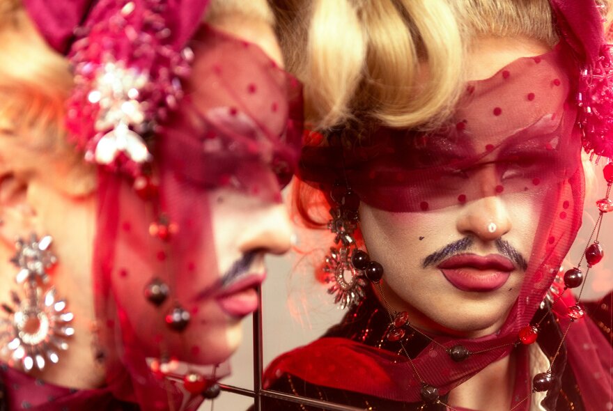 A close-up image of a drag artist looking into a mirror, wearing a red mesh veil that obscures the upper half of their face.
