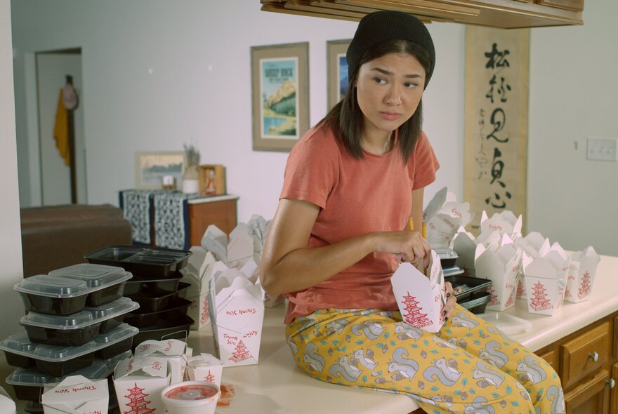 A still from the movie Egghead & Twinkie  showing a young woman sitting on a kitchen benchtop surrounded by Chinese takeaway containers.
