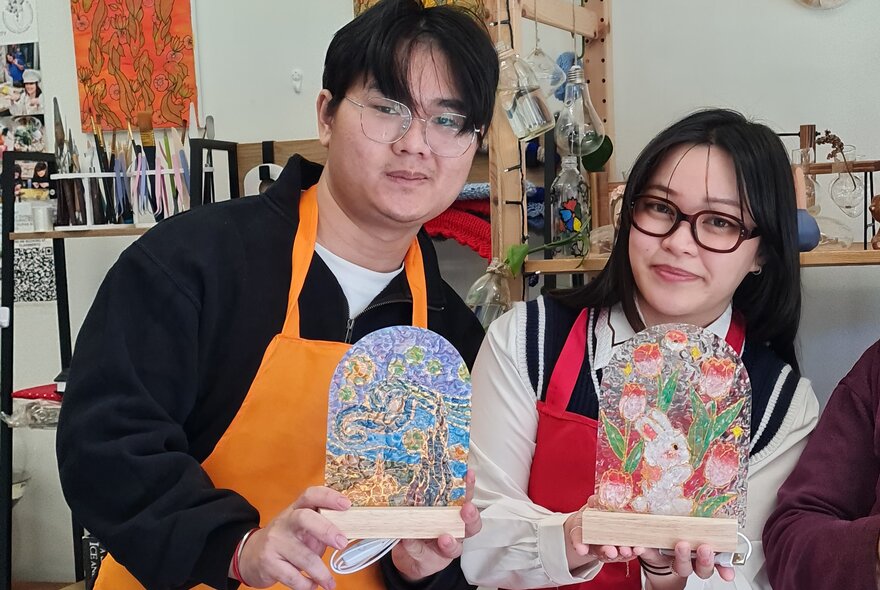 Two people in apron holding up hand painted glass lamps that they have made.