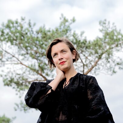 A woman, taken from below, looking contemplative with one hand on her neck and a tree behind her. 