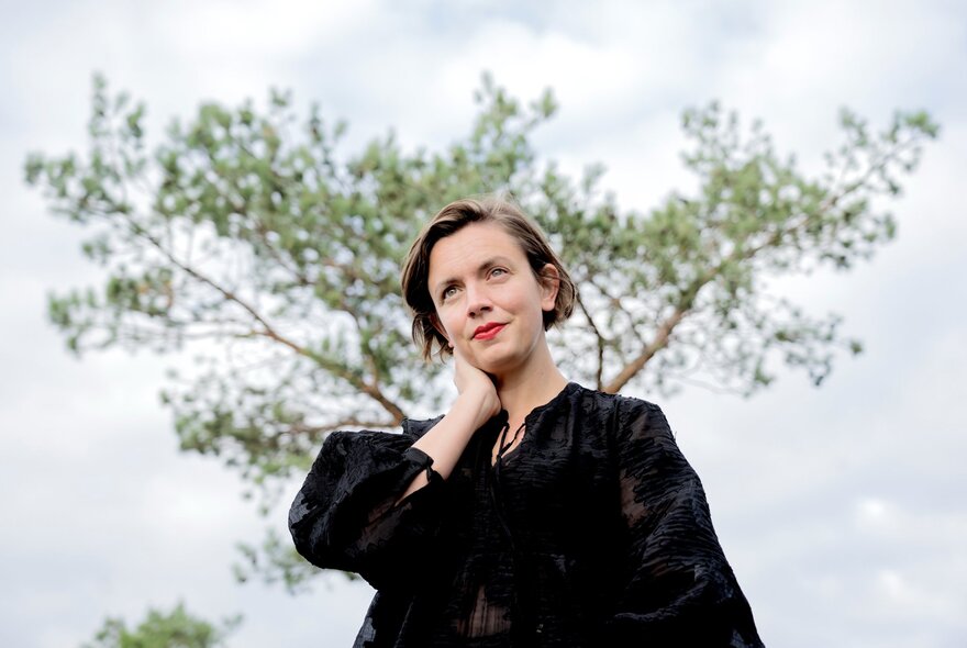 A woman, taken from below, looking contemplative with one hand on her neck and a tree behind her. 