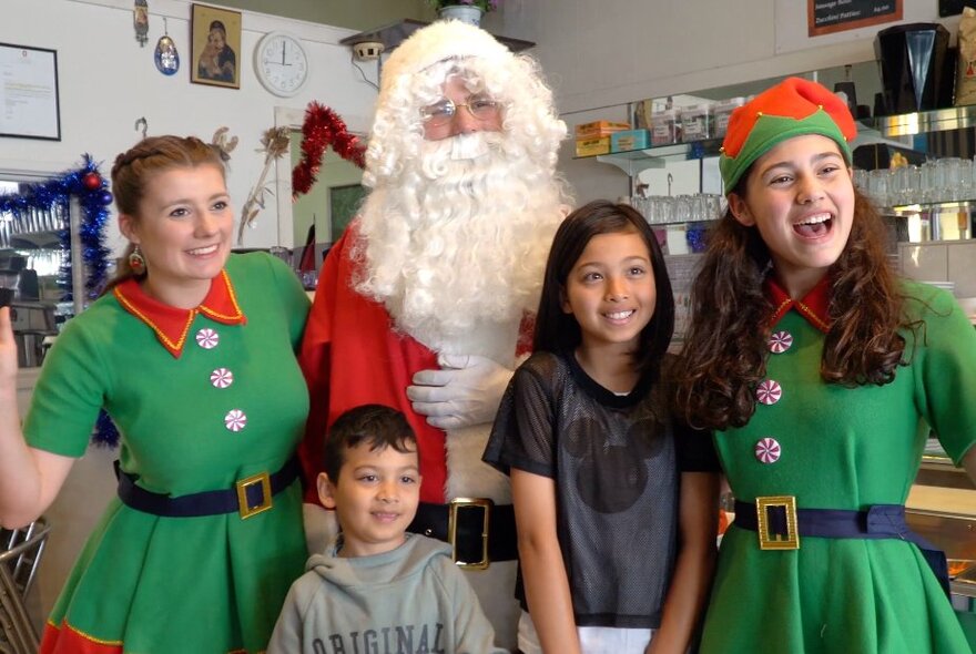 Two children with two Christmas elves wearing green and red tunis, with Santa in a cafe.