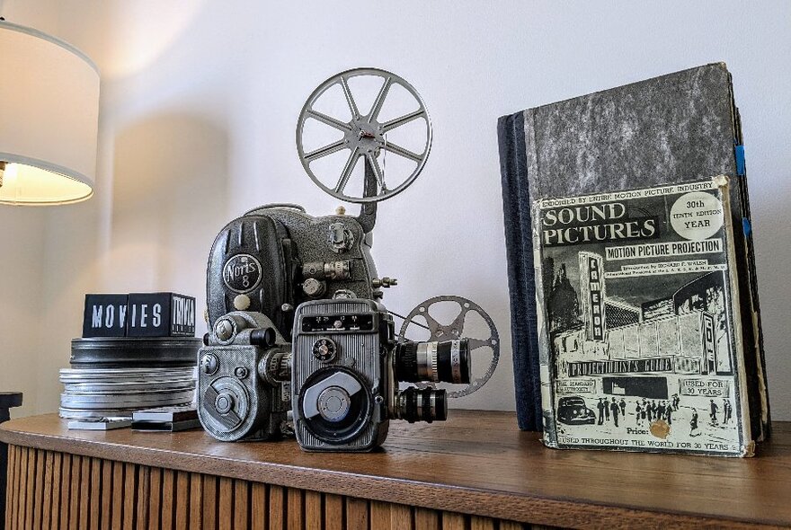 Old-fashioned film camera on glossy wooden surface, with movie reels to left and upright notebook to right.